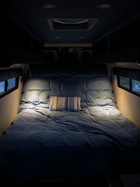 This RV rental's cozy bed features denim-blue bedding, a striped pillow, and bright reading lights for comfortable travels.