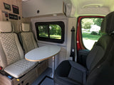 Interior of an RV rental: light-colored quilted seats, an oval table, control panels, and a view of a green yard.