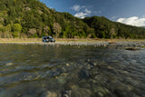 Grey RV rental parked on a rocky riverbank with clear water, backed by a large forested mountain.