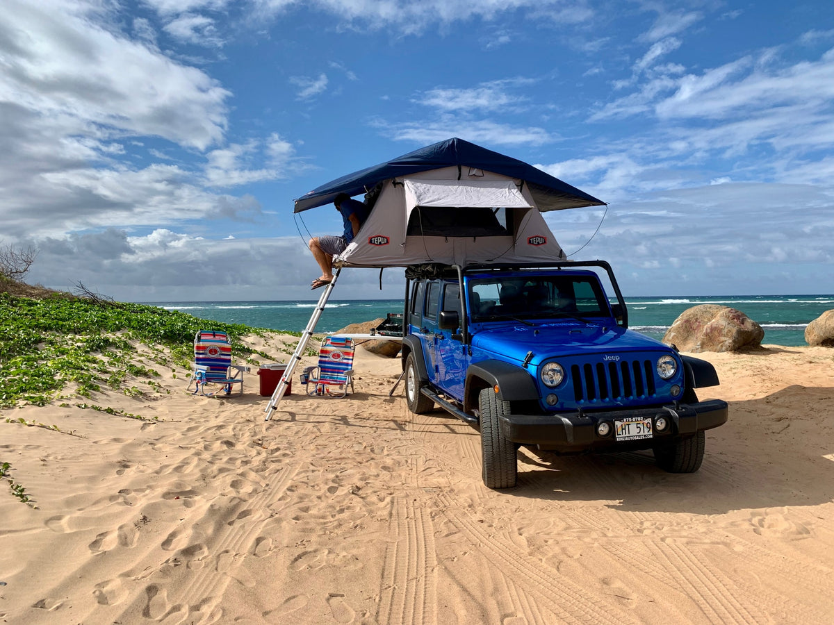 Blue Jeep with rooftop tent camping on a sandy beach. Perfect RV rental for ocean adventures. Chairs, cooler ready.