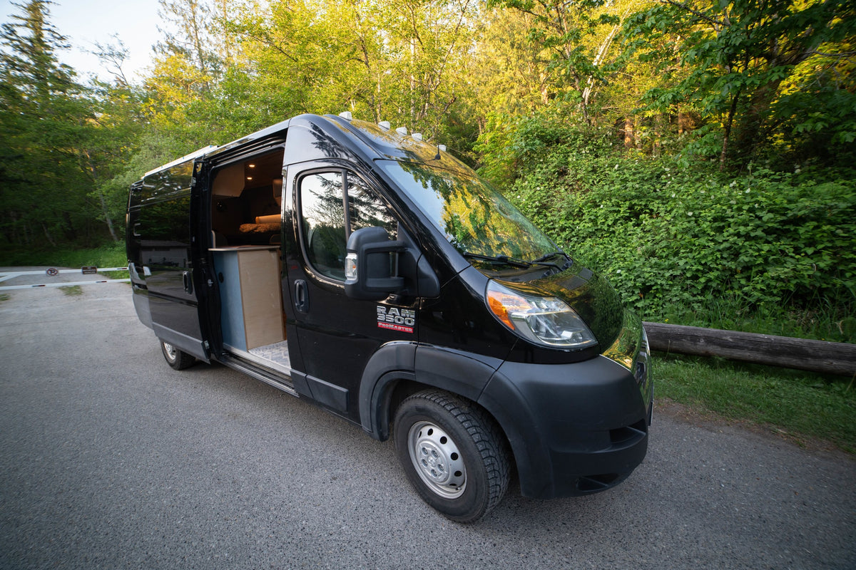 Black RV rental camper van parked on an asphalt road by a green forest. Its open side door reveals a cozy wooden interior. — photo 5