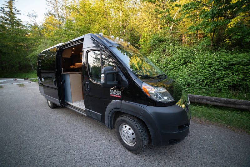 Black RV rental camper van parked on an asphalt road by a green forest. Its open side door reveals a cozy wooden interior. — photo 5