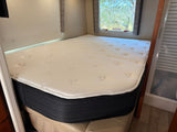Inside an RV rental: a plush, white mattress fills the cozy sleeping nook, with a window view of trees.