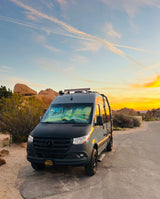 This custom Sprinter RV rental enjoys a vibrant desert sunset, parked amidst large rocks. Ideal for national park adventures.