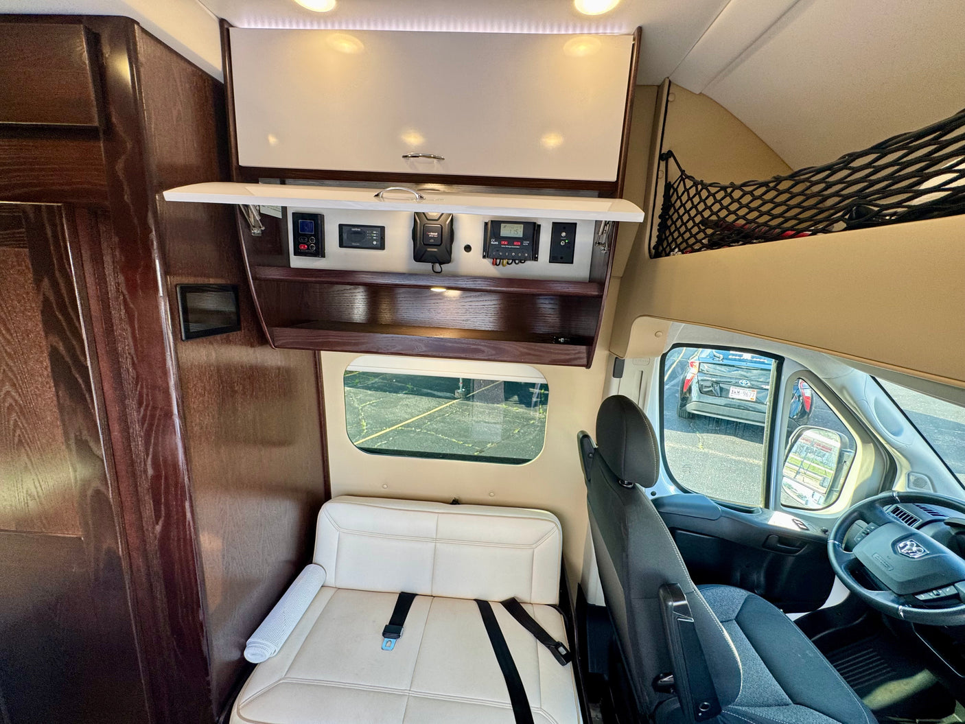RV rental van interior: control panels (solar, inverter), open overhead storage, passenger seating, and exterior view through windows.