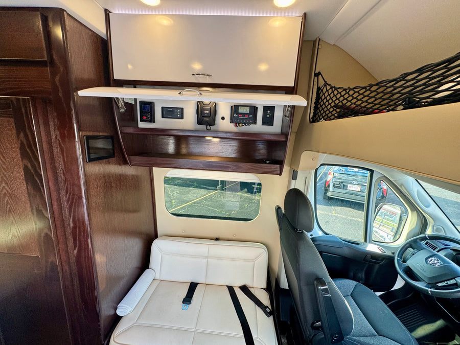 RV rental van interior: control panels (solar, inverter), open overhead storage, passenger seating, and exterior view through windows.