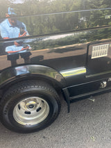 Close-up of a black RV rental's side, showing its tire, utility panel, and a clear reflection of the photographer.