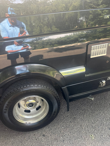Close-up of a black RV rental's side, showing its tire, utility panel, and a clear reflection of the photographer.