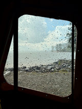 Cozy rainy day view from an RV rental window, with raindrops on glass, a body of water, and a shore beyond.