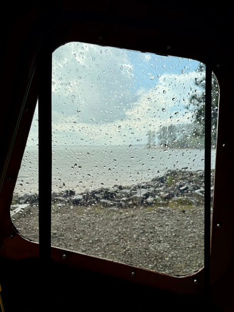 Cozy rainy day view from an RV rental window, with raindrops on glass, a body of water, and a shore beyond.