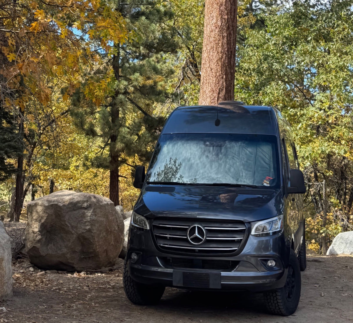 Black Mercedes Sprinter RV rental parked in a fall forest with yellow leaves and large rocks under a blue sky.|Compact RV rental interior featuring a bed