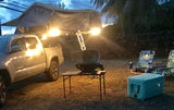 Night camping setup: Pickup truck with illuminated rooftop tent, grill, chairs, cooler.