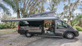 Grey camper van RV rental with extended pop-top roof and awning. Parked outside, showcasing its open interior and nature backdrop.