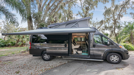 Grey camper van RV rental with extended pop-top roof and awning. Parked outside, showcasing its open interior and nature backdrop.