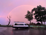 Stunning pink sunset and faint rainbow over an RV rental parked on a wet road, with dark trees and misty mountains.