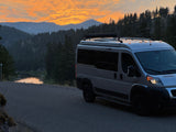 An RV rental camper van parked on a road at sunset, with a lake, mountains, and trees in the background.