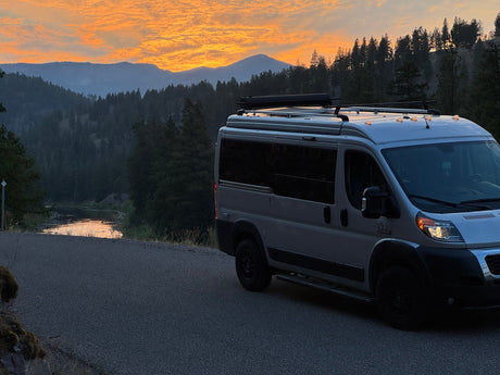 An RV rental camper van parked on a road at sunset, with a lake, mountains, and trees in the background.