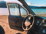 Inside a Ford rv rental, the driver's door is open to a scenic lakeside view. Sandy shore, blue water, and snowy mountains beyond.