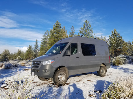 Grey Sprinter camper van **rv rental** in a snowy forest. Enjoy winter adventures with this off-road-ready vehicle.