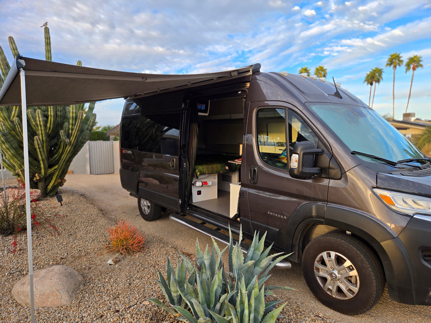 Entegra camper van rv rental with extended awning, parked in a desert landscape. Side door open, revealing interior, cacti & palm trees.