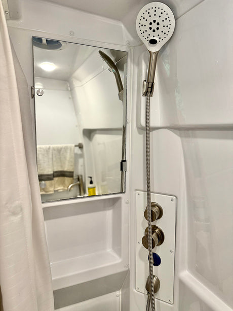 Compact RV rental shower: white stall with handheld showerhead, temperature controls, mirror, and shelf.