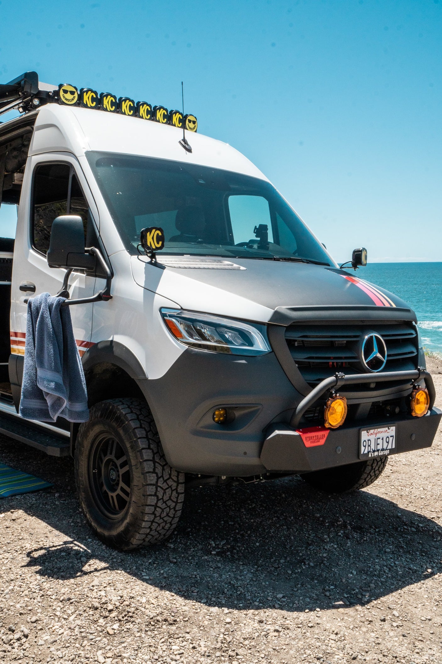 This custom Mercedes Sprinter overland RV rental, featuring off-road tires and KC lights, is parked by the ocean.