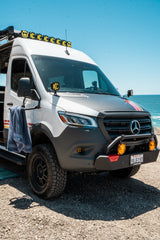 This custom Mercedes Sprinter overland RV rental, featuring off-road tires and KC lights, is parked by the ocean.