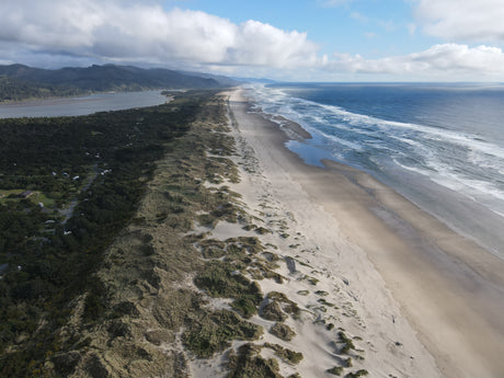 Aerial view: expansive sandy beach meeting ocean waves, backed by dunes, forest, river, and an RV rental campground.