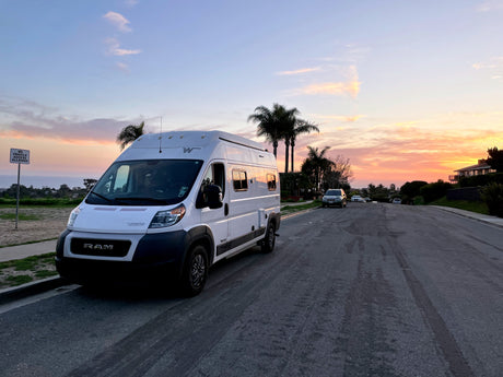 White RV rental parked on a coastal road at sunset, with palm trees and ocean views. Explore van life adventures!