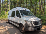 White Freightliner camper van in a sun-dappled forest. Perfect for an RV rental adventure, featuring a roof-mounted light bar.