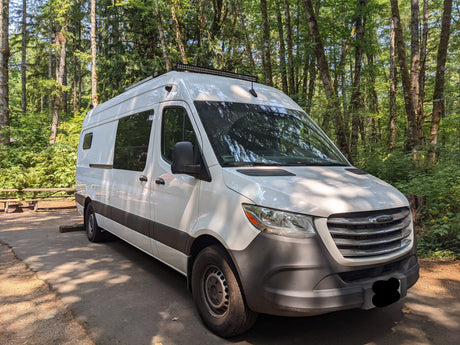White Freightliner camper van in a sun-dappled forest. Perfect for an RV rental adventure, featuring a roof-mounted light bar.