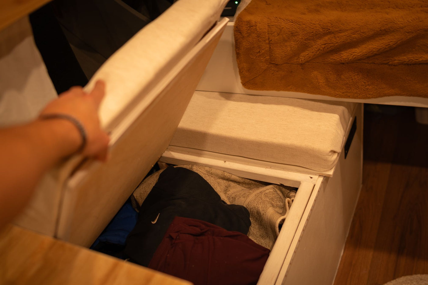 Under-bench storage in an RV rental. A hand lifts the cushion to reveal folded clothes inside, perfect for travel organization.
