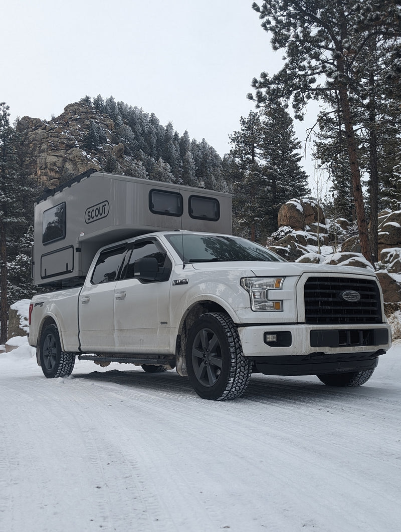White Ford F-150 with a grey Scout truck camper on a snowy mountain road with pine trees. An ideal RV rental for winter camping. — photo 1