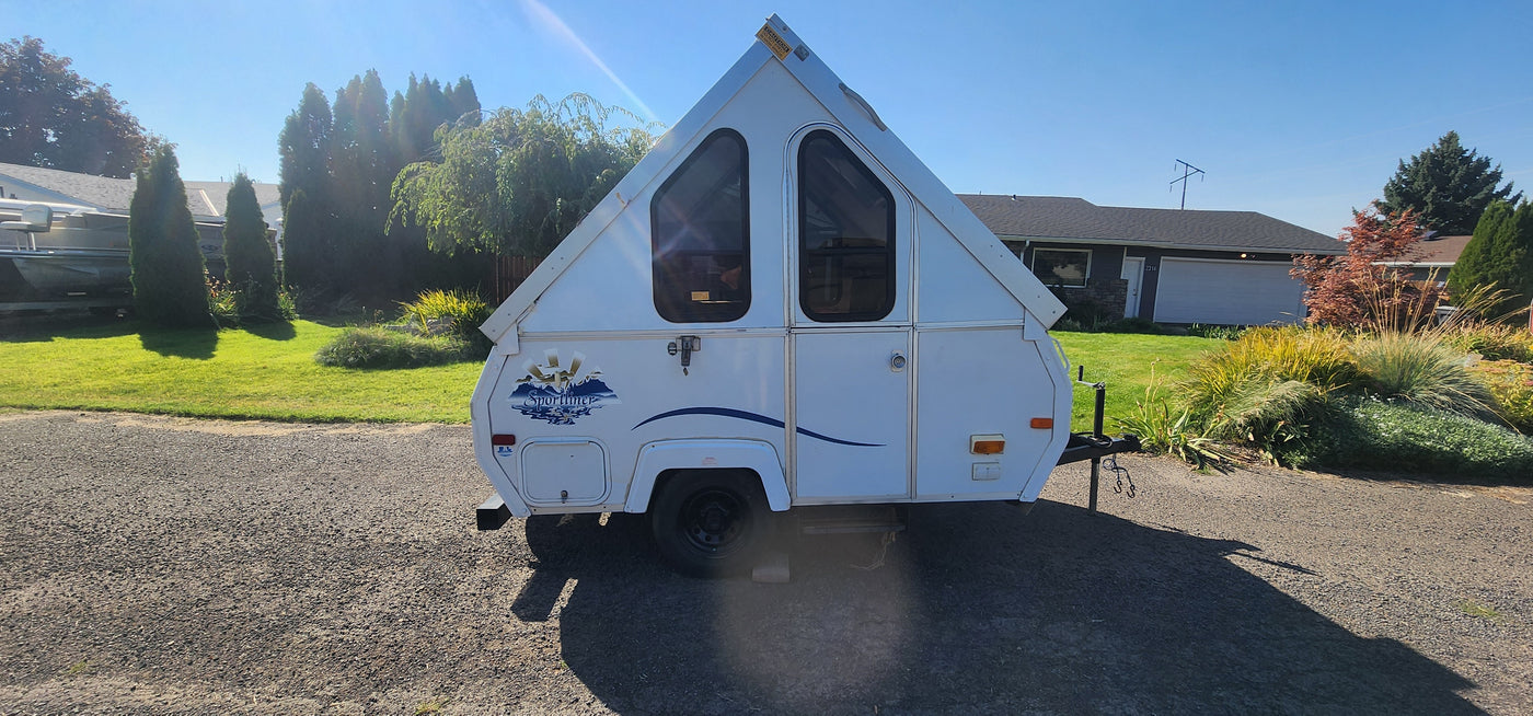 White A-frame rv rental camper with triangular windows, parked on gravel in a sunny residential setting.