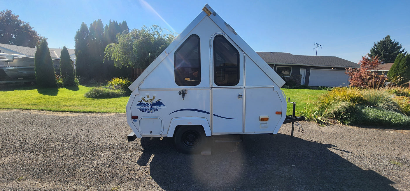 White A-frame rv rental camper with triangular windows, parked on gravel in a sunny residential setting.