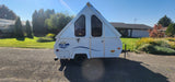 White A-frame rv rental camper with triangular windows, parked on gravel in a sunny residential setting.