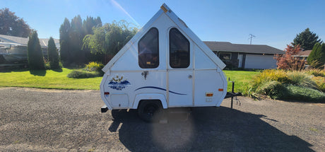 White A-frame rv rental camper with triangular windows, parked on gravel in a sunny residential setting.