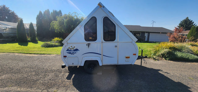White A-frame rv rental camper with triangular windows, parked on gravel in a sunny residential setting.
