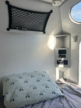 Cozy rv rental interior: bed with patterned pillow, overhead net, power panel, & round window to blue sky.