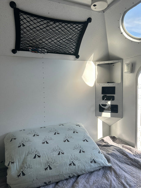 Cozy rv rental interior: bed with patterned pillow, overhead net, power panel, & round window to blue sky.