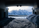 Inside an RV rental, a comfy bed overlooks a serene snow-covered landscape & majestic Teton mountains. Ultimate winter camp view.