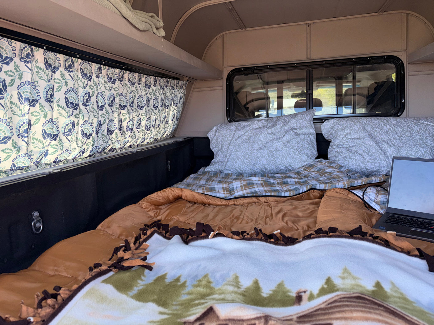 Cozy rv rental interior with a bed, laptop, pillows, and floral curtains, ready for a comfortable camping trip.