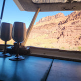 From an RV rental, two stemless wine glasses frame a view of a stunning red rock canyon under a clear blue sky. Serene desert landscape.