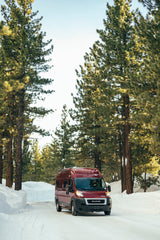 A red RV rental van drives on a snowy road through a pine forest, perfect for winter adventures.