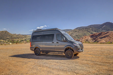 Off-road RV rental: A rugged Mercedes Sprinter adventure van in a scenic mountain landscape with red rocks, parked on a dirt lot.