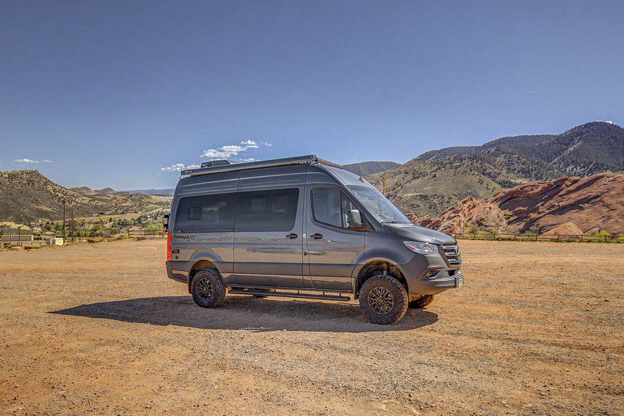 Off-road RV rental: A rugged Mercedes Sprinter adventure van in a scenic mountain landscape with red rocks, parked on a dirt lot.
