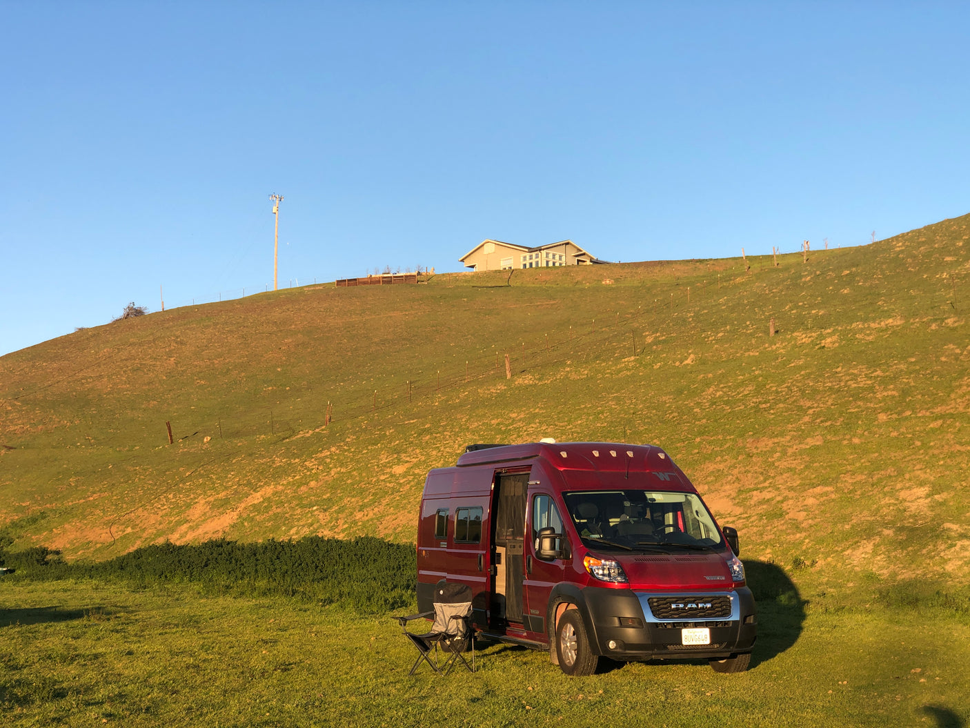 A red **rv rental** camper van is set up for outdoor relaxation on a sunny green field, with a hill and house behind.