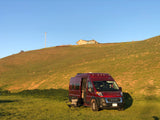 A red **rv rental** camper van is set up for outdoor relaxation on a sunny green field, with a hill and house behind.