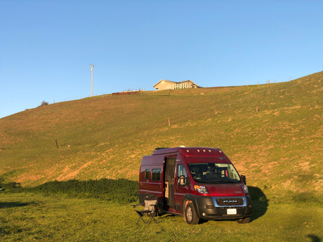 A red **rv rental** camper van is set up for outdoor relaxation on a sunny green field, with a hill and house behind.