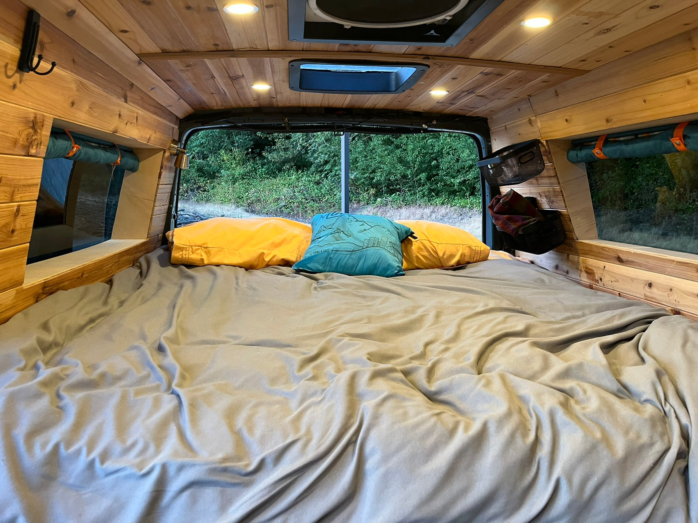 Inside a cozy RV rental: Wood-paneled camper van bedroom with a comfy bed, colorful pillows, and a scenic view of lush trees. — photo 4